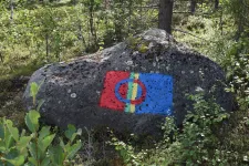 En sten omgiven av grönska med den samiska flaggan målad på. Foto.